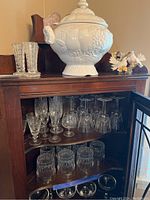White ceramic soup tureen on top of wood cabinet alongside two white ceramic bird figurines. Inside cabinet are multiple types of glassware arranged on shelves.