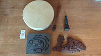 Lot displayed showing hand drum, drumstick, art tile, and carved figurine on wooden surface