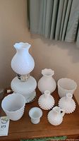 Full view of all eight milk glass items on table including the lamp and seven vases or containers, all hobnail pattern.