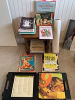 Photo of the assortment of music books stacked on and around a wooden side table with a rolled white sheet beside it.