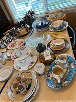 Overview of entire lot showing 3-tier floral pastry tray, assorted plates, bowls, glass platters, trivets, and cutlery on table.