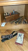 Photo showing multiple vintage scissors, wooden box, two leather belts, manual hair clipper in red box on wood floor