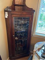 Full-height view of solid wood corner cabinet in room corner, glass-paneled door closed, showing overall size and style.