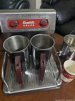 Front view of Campbell's Soup warmer showing two metal mugs with maroon handles inside warming tray. Red brand label and control knobs visible.