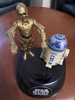C3PO and R2D2 banks on black oval base with Star Wars logo in white, shown on table.