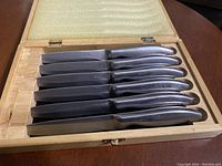 Photo of six steak knives arranged inside a foam-lined wooden box showing blades and handles.