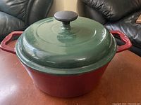 Red cast iron Dutch oven with green lid on a brown surface, showing overall exterior with handles and knob.