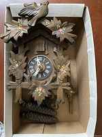 Top view of a traditional wooden cuckoo clock placed in a cardboard box showing carved leaves, bird on top, clock face with Roman numerals and two pine cone weights.