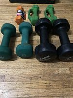 Front view of seven dumbbells arranged on a wooden surface. Includes different colors and weights with visible coatings and brand markings.