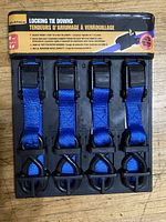 Set of 5 brand new locking tie downs with blue straps in original packaging shown from top view.