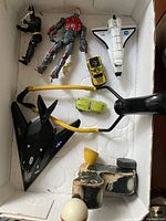 Top view of box containing black Batman-like action figure, red and silver robot figure, two small toy cars (yellow and lime green), sling shot planes, wooden pull duck toy, wooden wind-up car, and black wind-up key.