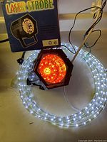Photo showing coiled white LED rope light beside a black hexagonal laser strobe light emitting red light, and the laser strobe light box in the background.