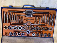 Top-down view of tap and die set in open metal case with wooden insert, showing taps, dies, and related holders.