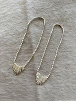 Two white mother of pearl beaded necklaces laid flat on a fabric background, showing entire length with fan-shaped pendant detail.