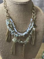 Clear front view of a vintage 5 strand necklace on a beige fabric bust showing details of the glass beads, crystal beads, irregular stones, and gold tassels.