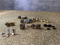Photo showing all cufflinks arranged in two rows on fabric background, displaying a variety of styles and colors.