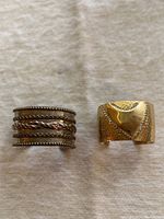 Two vintage cuff bracelets, one silver-tone multi-band and one brass with a heart design, displayed together on a fabric background.
