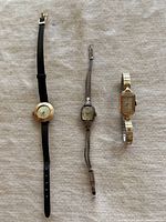 Three vintage gold filled watches laid flat on a light fabric surface: one with round bezel and black leather strap, one with intricate chain bracelet, and one with rectangular face and stretch band.