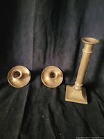 Photo of three brass candle holders on black background: two short round with wide drip trays and one tall with square base and column stem