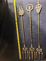 Three vintage brass forks with detailed decorative handles and four-pronged heads, shown beside a measuring tape for scale.