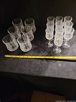 Front view of 6 crystal tumblers and 10 small stemmed crystal glasses arranged with a measuring tape showing sizes.