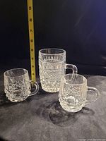 Three vintage crystal mugs with textured patterns and handles on a black surface with measuring tape showing sizes