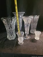 Six clear crystal vases displayed on black background with measuring tape showing height for scale