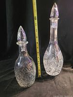 Two vintage cut glass decanters standing side by side against a black background with a measuring tape showing height.