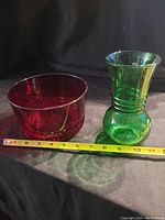 Red glass bowl and green glass vase placed next to a measuring tape for size reference on a black background.