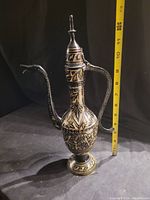 Front angle of vintage metal teapot with visible engraved floral and geometric patterns, measuring about 12 inches tall with a yellow measuring tape beside it.