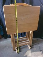 Photo of 4 folded wooden TV tables standing upright with measuring tape for height reference (about 27-28 inches tall).