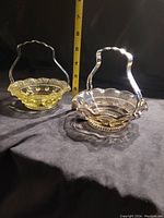 Front and side view of two vintage glass dishes with metal handles on a dark cloth background with a measuring tape showing size.