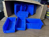 Sixteen blue Akro plastic bins arranged with some inside a cardboard box on a tabletop, showing front access cutout and shape.