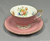 Close-up of Aynsley teacup with pink exterior and floral inner design, gold rim and handle, paired with matching pink saucer with gold rim.