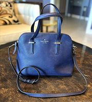 Front view of blue leather Kate Spade purse with double handles and gold-tone logo, sitting on a table.