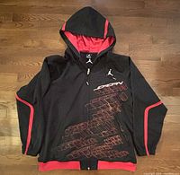 Front view of Nike Air Jordan hooded jacket showing zipper, graphic design with Jordan text and Jumpman logo, red accents on black fabric.