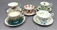 Four Royal Albert teacup and saucer sets with varied floral patterns and gold trim on grey background