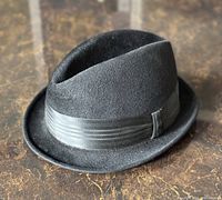 Black felt men's fedora hat with grosgrain ribbon shown angled from front and side on a brown surface.