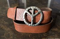 Close-up photo showing brown textured leather belt rolled with circular metal peace symbol buckle with patina