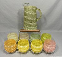Full set with pitcher and all 8 glasses in metal/wood rack showing the pitcher in the back and 8 cups arranged front and back in the rack.