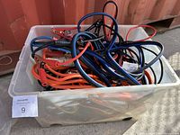 Clear rectangular plastic bin filled with bundled booster cables of various colors including red, blue, orange, and black. Clamps visible on several cables.