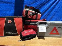 Four emergency kits arranged on blue cloth background including three fabric bags (one labeled Auto Safety Kit) and one grey plastic case with an emergency triangle inside.