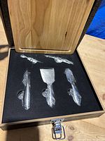 Opened wooden gift box showing four stainless steel cheese cutting tools resting in fitted black foam alongside a wooden cutting board above them.
