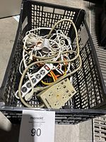 Top-down photo showing mixed pile of power strips and extension cords in a black plastic crate, some cords tangled.