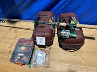 View of two maroon hard plastic tackle boxes, one fishing rod with green reel, and a smaller plastic tackle box with bright floats and hooks plus resealable bag of hooks and lures.