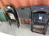 Group of folded black metal and plastic folding chairs stacked against a wall, showing various styles and conditions.