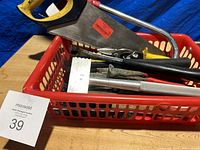 Photo showing assorted hand tools including hand saw, locking pliers, hammer, Stanley pliers, and a metal mallet in a red plastic basket.