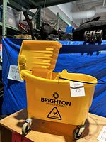 Yellow wheeled industrial mop bucket with Brighton logo, visible caster wheels, and caution graphic.