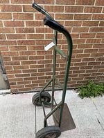 Front angle view of the green metal dolly showing one handle and a single wheel, against a brick wall.