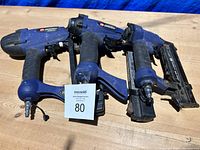 Lot of three Campbell Hausfeld blue pneumatic nailer tools, aligned on wooden surface, showing overall condition and size.